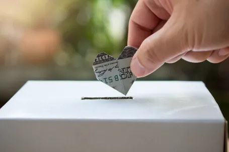 A dollar folded into a heart shape being dropped into a donation box