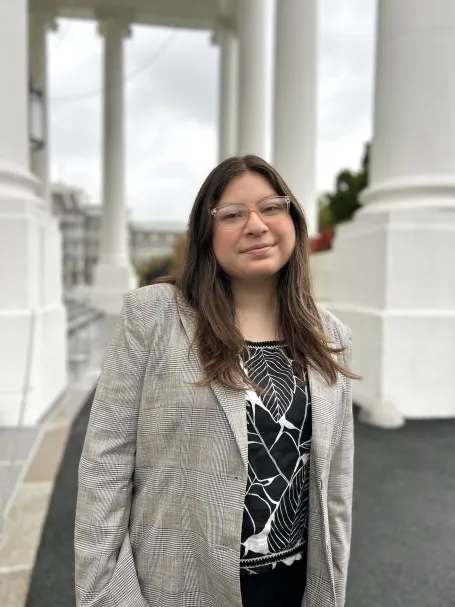 A photo of a woman with long brown hair and glasses outside a government building