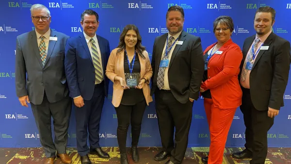 A woman wearing a tan blazer holds an award while flanked by IEA leadership