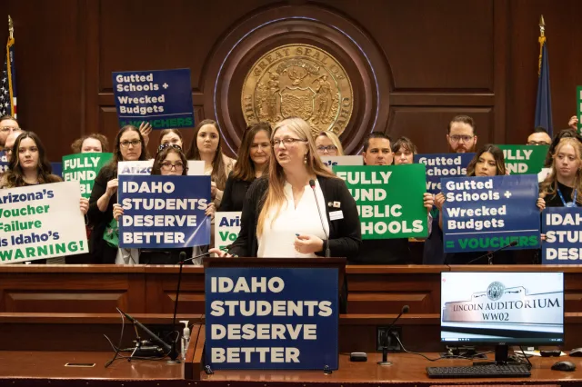 A woman speaks at a podium as a group of people hold blue, green and white anti-voucher posters