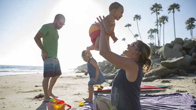 A family plays on a beach: A woman holds a toddler in the air as they laugh, and a man and young girl look at their sand castle tools together.