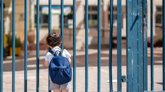 Small child wearing a blue backpack stands at locked gate.