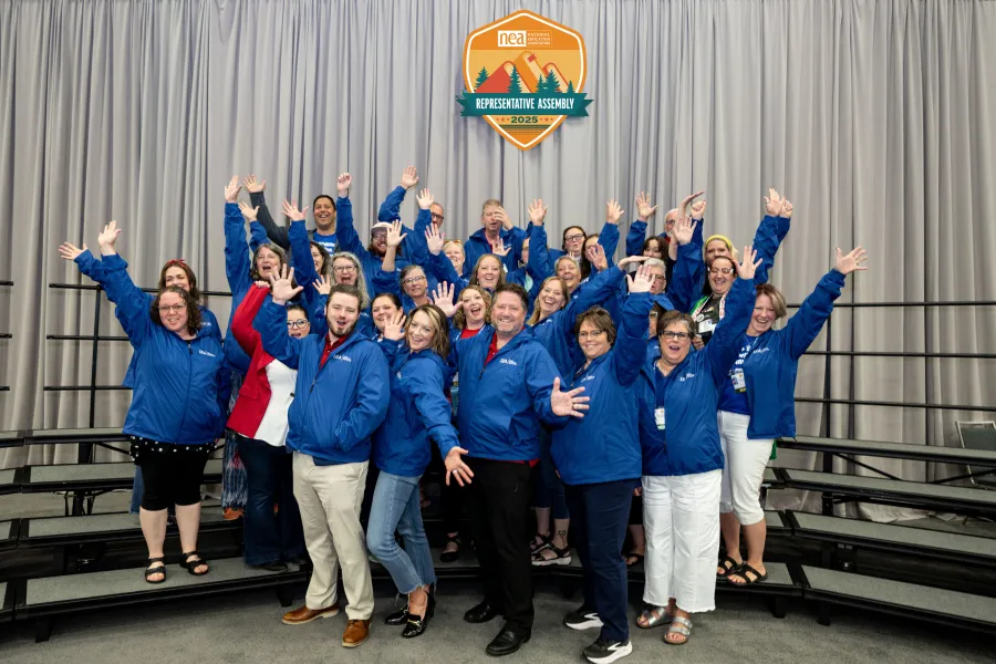 IEA's 2025 NEA Representative Assembly delegates pose for a delegation photo under the RA logo with their arms in the air, dressed all in blue.